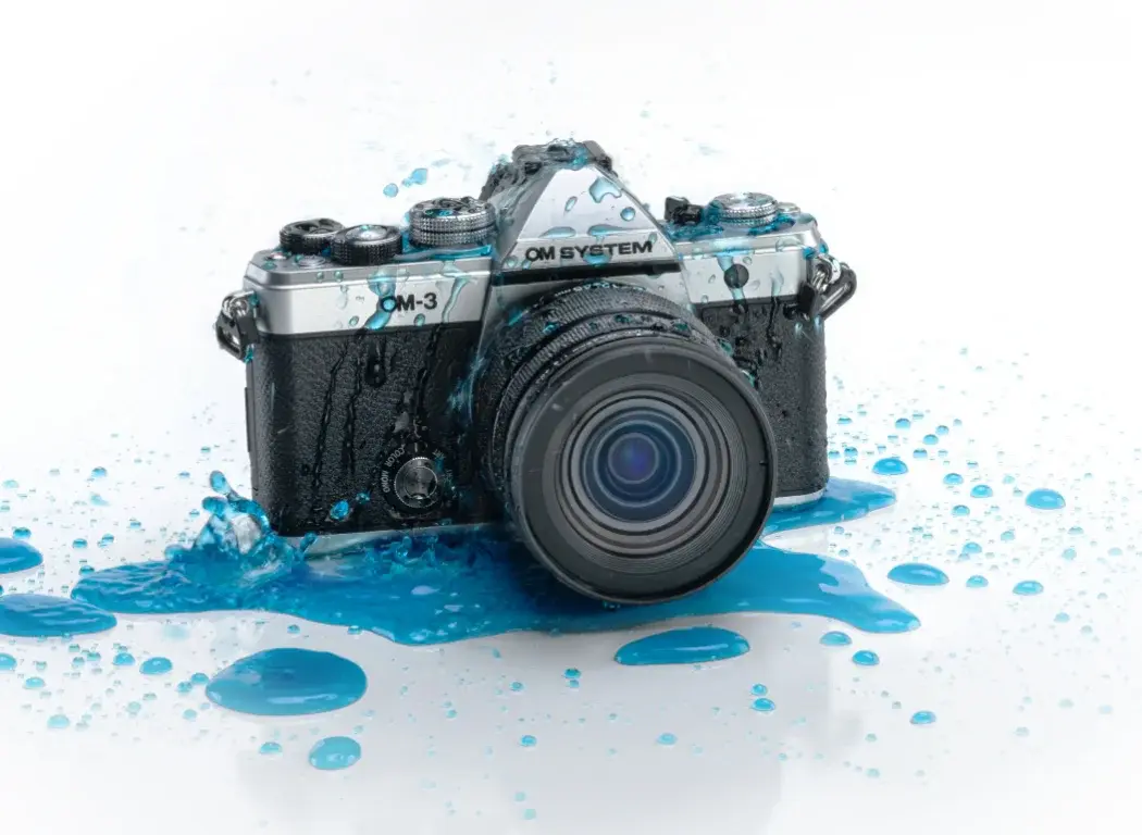 OM SYSTEM OM-3 camera sitting on a white table with water splashed all over it.